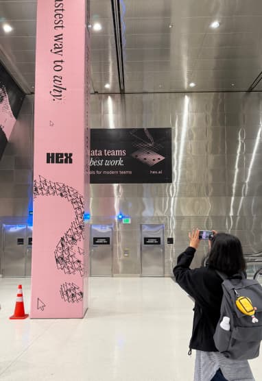 Person photographing a Hex out-of-home ad — a tall pink column with "fastest way to apply" and Hex branding — in an airport or transit hall