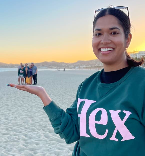 Hex team member in a green Hex sweatshirt on a beach at sunset, playfully holding tiny teammates in her outstretched palm